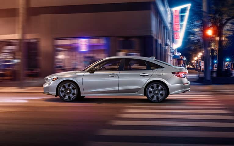 2027 Honda Civic Sedan Sport Touring Hybrid in Solar Silver Metallic side profile driving on a city street (Compact Sedan)